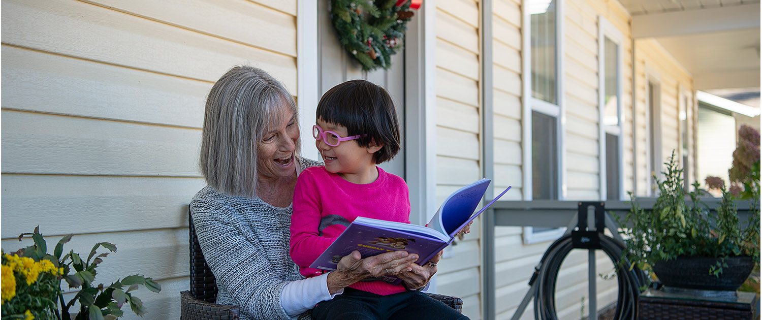 A child sits in a woman's lap on a porch while reading a book