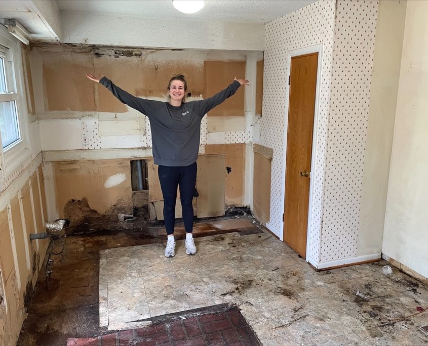 Natalie stands in the completely empty kitchen, having removed the cabinets, countertops, sink and stove.