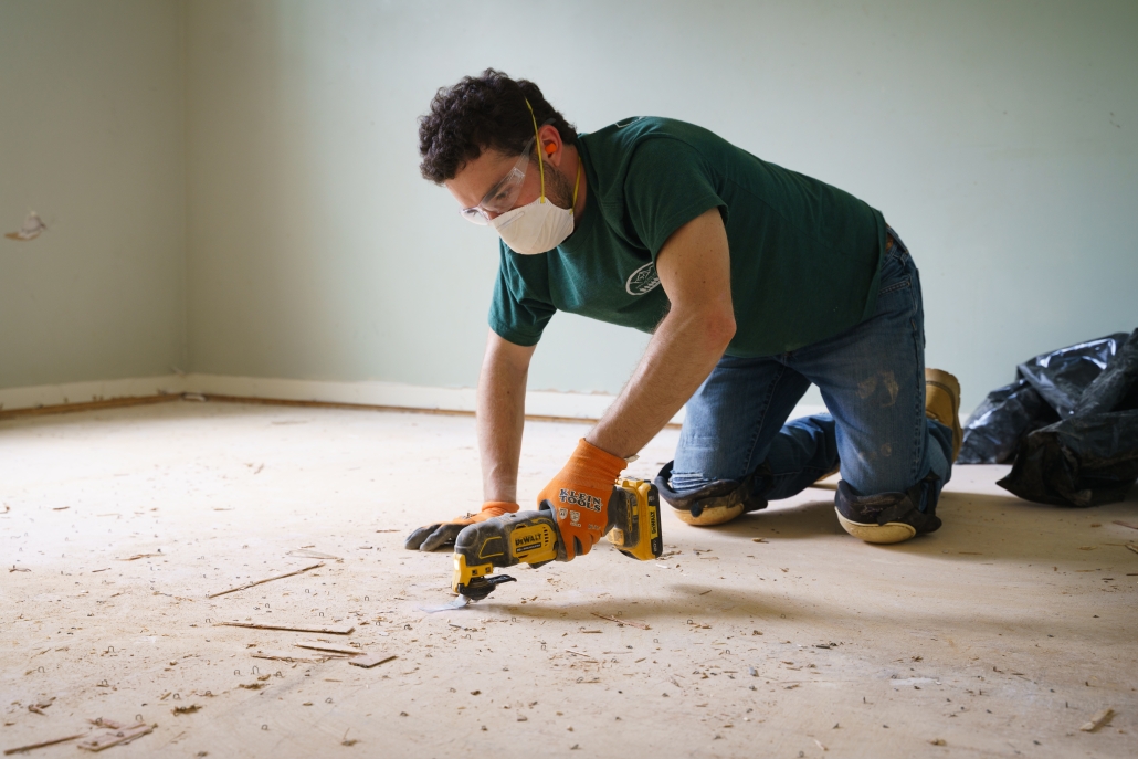 A volunteer uses a multitool while kneeling inside of a home in the middle of a repair. The subfloor of the home is exposed, and the volunteer is cutting out raised parts of the subfloor.