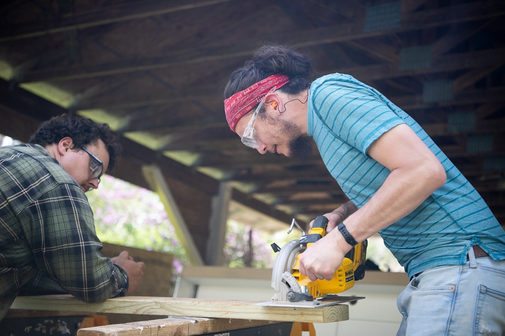 Two volunteers wearing safety glasses work with lumber under a covered construction area. One volunteer operates a circular saw to cut a wooden board supported by sawhorses while another volunteer steadies the material. Natural light filters through the open structure, highlighting the active construction workspace.