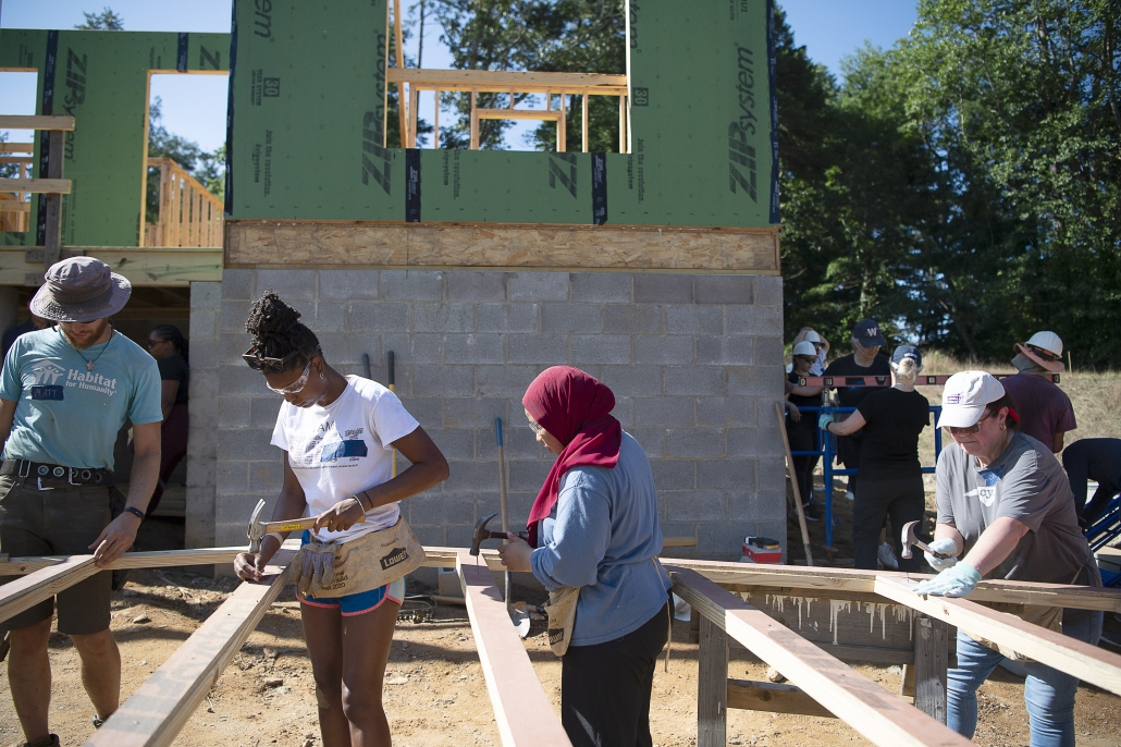 Volunteers assemble wooden wall frames at an active Habitat for Humanity construction site. Several people wearing safety glasses and tool belts use hammers to secure lumber, while a partially built house with concrete block foundation and green exterior sheathing stands in the background. Additional volunteers work nearby under a clear sky.