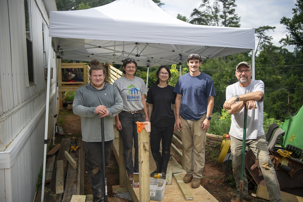 A group of volunteers stand with Construction Associate Audge under a pop-up tent just outside of a mobile home, where they have been working on repairs.