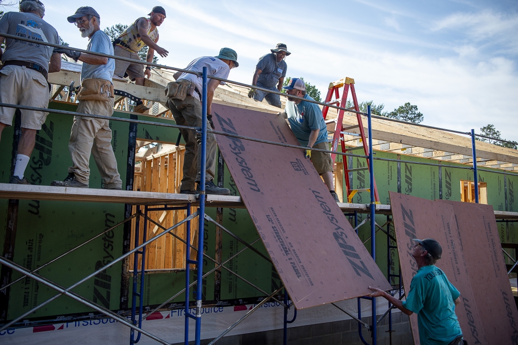 Volunteers working on scaffolding install roof sheathing on a Habitat for Humanity home under construction. Several people coordinate to lift and position large sheathing panels along the wall while others guide the materials from below. The partially framed house, scaffolding, and construction tools are visible beneath a bright sky.