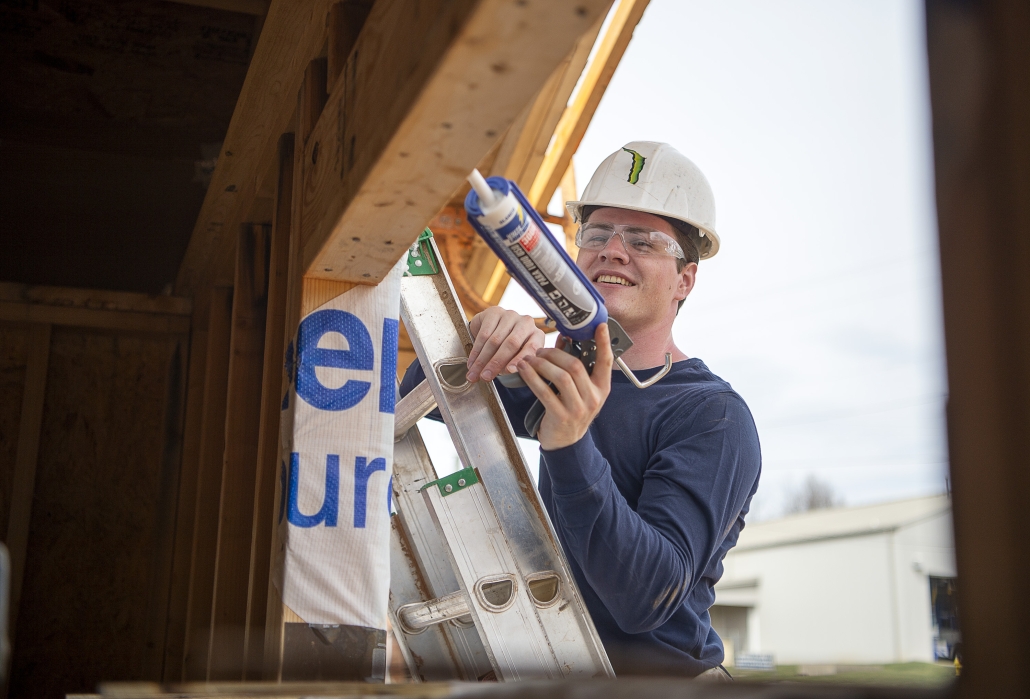 A volunteer wearing a hard hat and safety glasses applies sealant around a framed window opening while standing on a ladder outside a Habitat for Humanity home under construction. Exposed wood framing is visible around the work area.